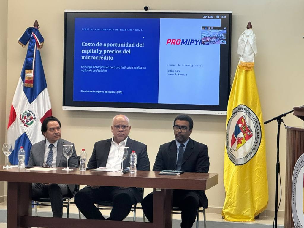 Tres hombres sentados en una mesa frente a una presentación en pantalla sobre el costo de oportunidad del capital y precios del microcrédito, con banderas y un sello de la universidad al fondo.