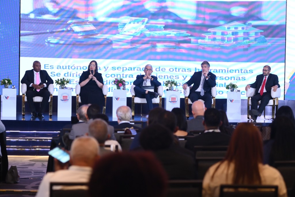 Panel de discusión con cinco ponentes en un evento, enfocado en temas legales y sociales. Pantalla de fondo con texto y gráficos relacionados.