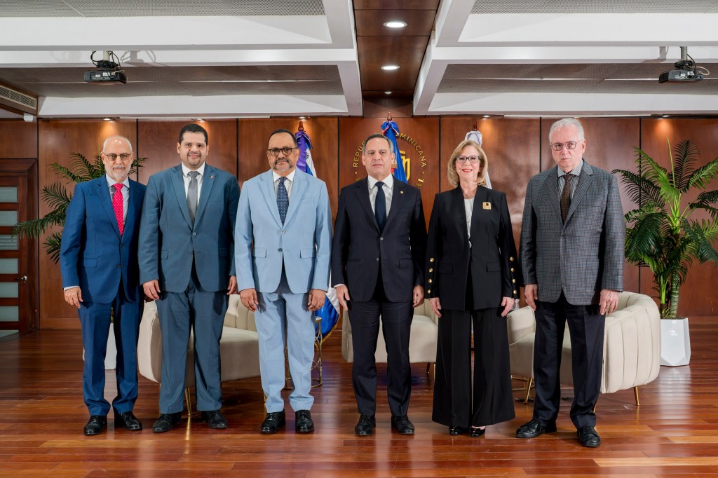 Grupo de seis personas posando para una foto en un ambiente formal, con fondo de madera y plantas decorativas.