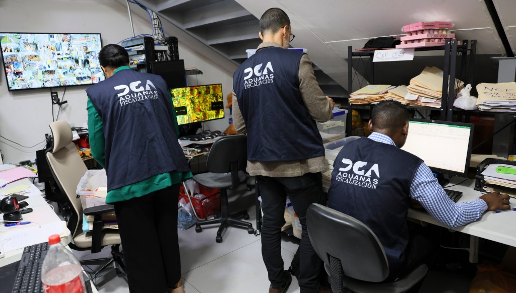 Tres empleados de aduanas trabajan en una oficina, uno de pie revisando documentos y dos sentados frente a computadoras. Pantallas con imágenes en el fondo.