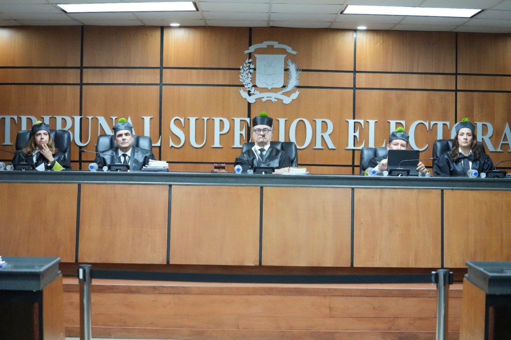 Jueces en una sala de tribunal, vistiendo togas y gorros, frente a un panel con el texto 'TRIBUNAL SUPERIOR ELECTORAL'.