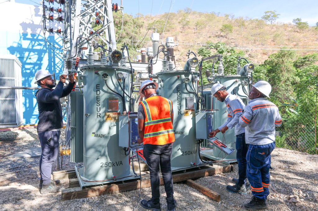 Trabajadores técnicos ajustando transformadores eléctricos en una estación de energía, con vegetación al fondo.
