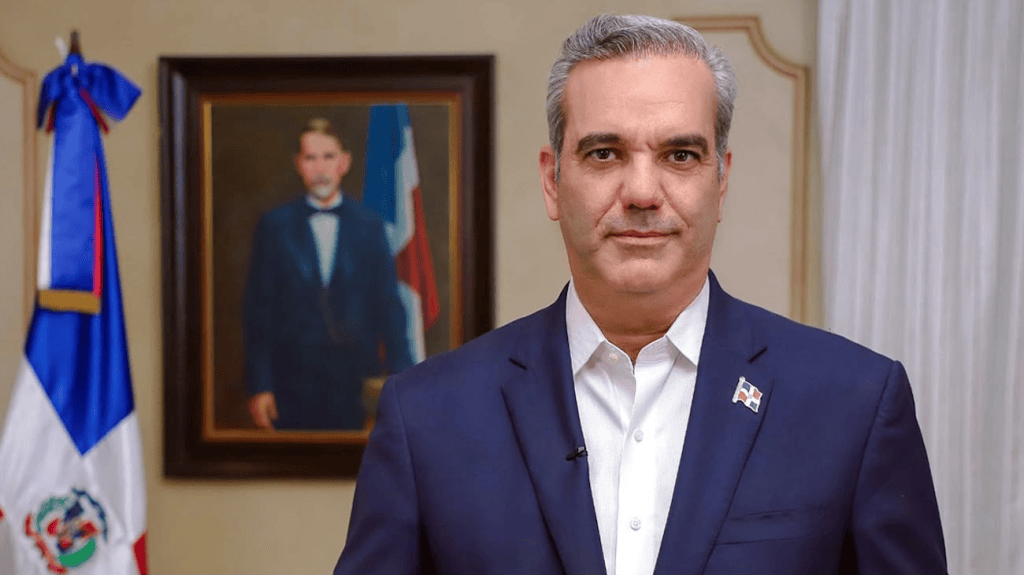 Hombre en traje oscuro frente a un retrato enmarcado y una bandera de la República Dominicana de fondo.