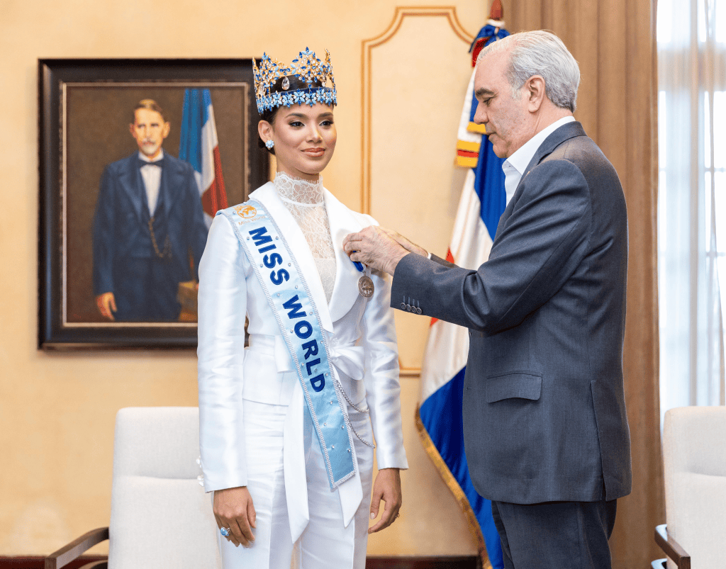 Una mujer con la corona de Miss Mundo, vestida de blanco, recibe una medalla de un hombre mayor en un entorno elegante, con un retrato y banderas de fondo.