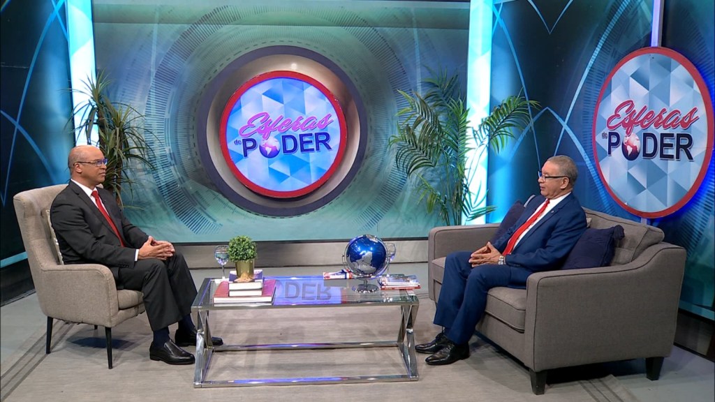Dos hombres sentados en un set de televisión durante un programa titulado 'Esferas de Poder'. Uno lleva un traje oscuro y el otro un blazer claro, con una mesa de cristal entre ellos y decoración de plantas.