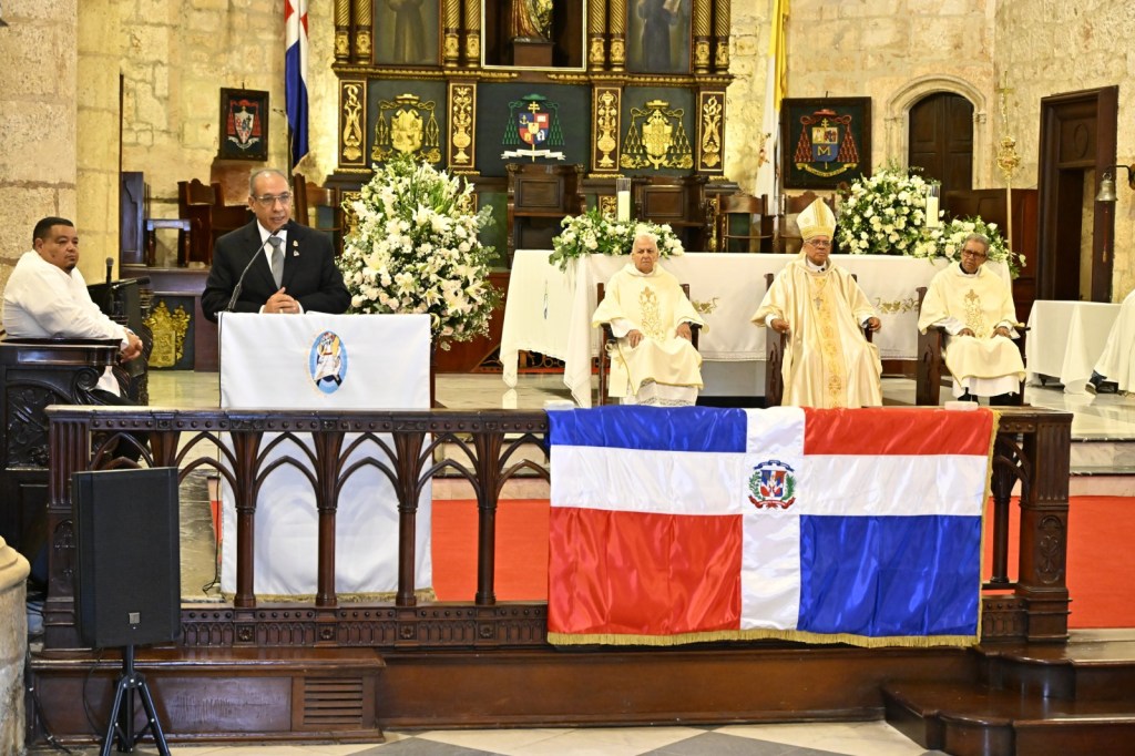 Un hombre en traje de gala habla en el altar de una iglesia, mientras otros tres hombres vestidos con vestimenta eclesiástica lo escuchan. Hay una bandera dominicana en primer plano y arreglos florales decoran el entorno.