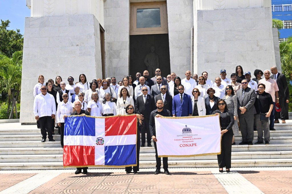Grupo de personas posando frente a un monumento, sosteniendo una bandera dominicana y una pancarta del Consejo Nacional de la Persona Envejeciente (CONAPE).