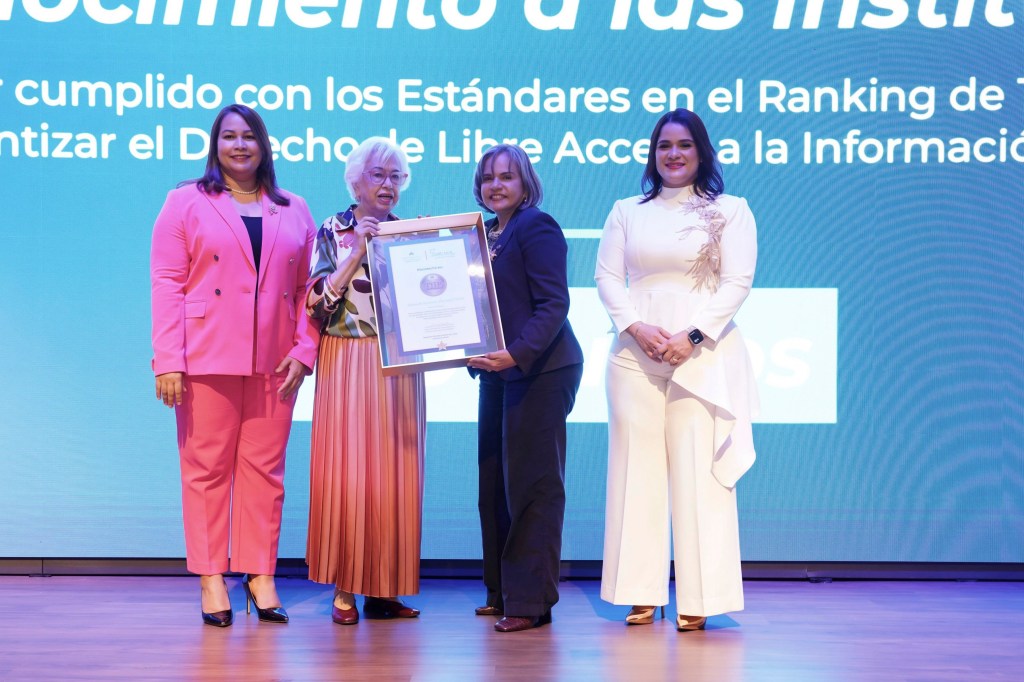 Cuatro mujeres sonriendo mientras sostienen un certificado en un escenario, con un fondo que destaca un mensaje sobre el cumplimiento de estándares relacionados con el acceso a la información.