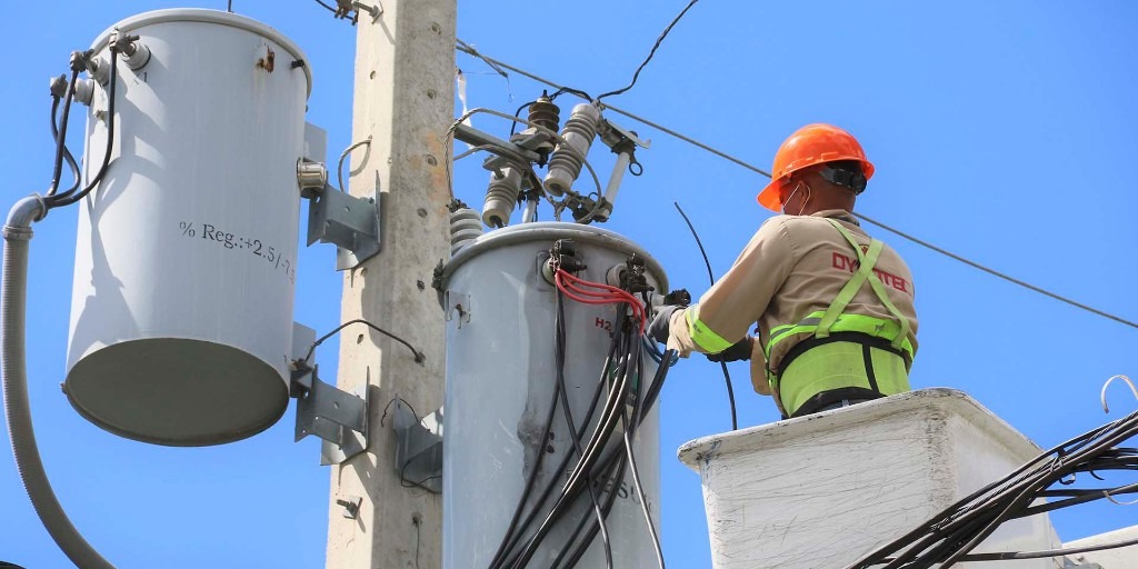 Trabajador de electricidad realizando mantenimiento en transformador en un poste, usando casco naranja y arnés de seguridad, con un cielo azul al fondo.