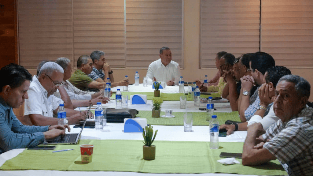 Reunión en una mesa larga con participantes sentados, discutiendo en un ambiente formal.