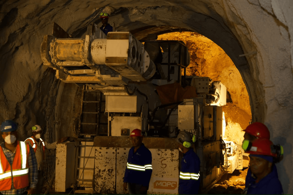 Operarios en un túnel de minería, con maquinaria pesada y pared de roca iluminada al fondo.