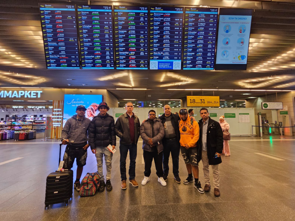 Grupo de hombres en un aeropuerto, frente a un panel de llegadas y salidas con horarios de vuelos. Algunos llevan equipaje.