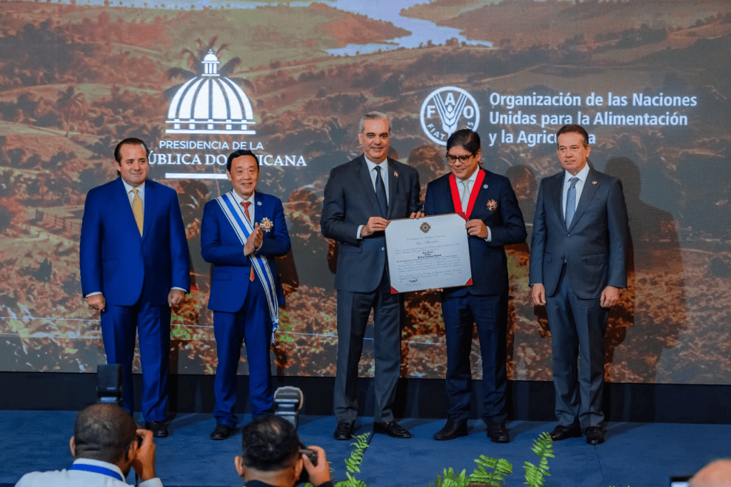 Ceremonia de entrega de reconocimiento en la República Dominicana, con varios asistentes, incluyendo dignatarios y un homenajeado que sostiene un certificado.