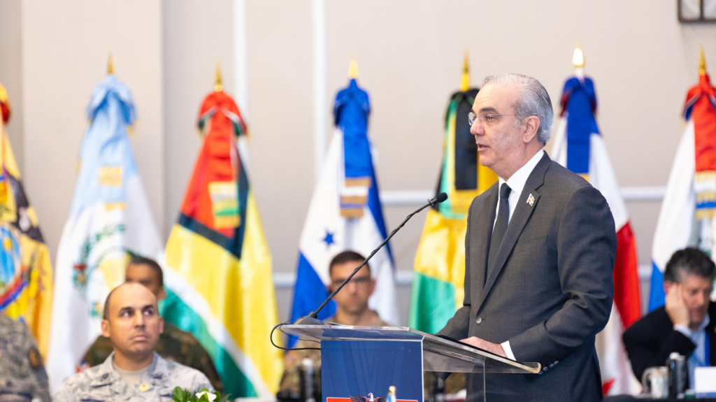 Un hombre de pie detrás de un podio, hablando en un evento formal, con banderas de diferentes países en el fondo y asistentes en la audiencia.