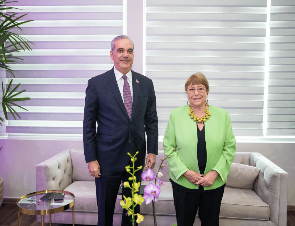 Un hombre y una mujer de pie en una sala bien iluminada, junto a un sofá claro. El hombre lleva un traje oscuro y la mujer viste un abrigo verde claro con un collar llamativo. Hay plantas y orquídeas en el entorno.