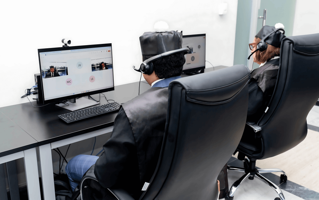 Dos personas con togas y auriculares participan en una videoconferencia en sus computadoras en un entorno de oficina.