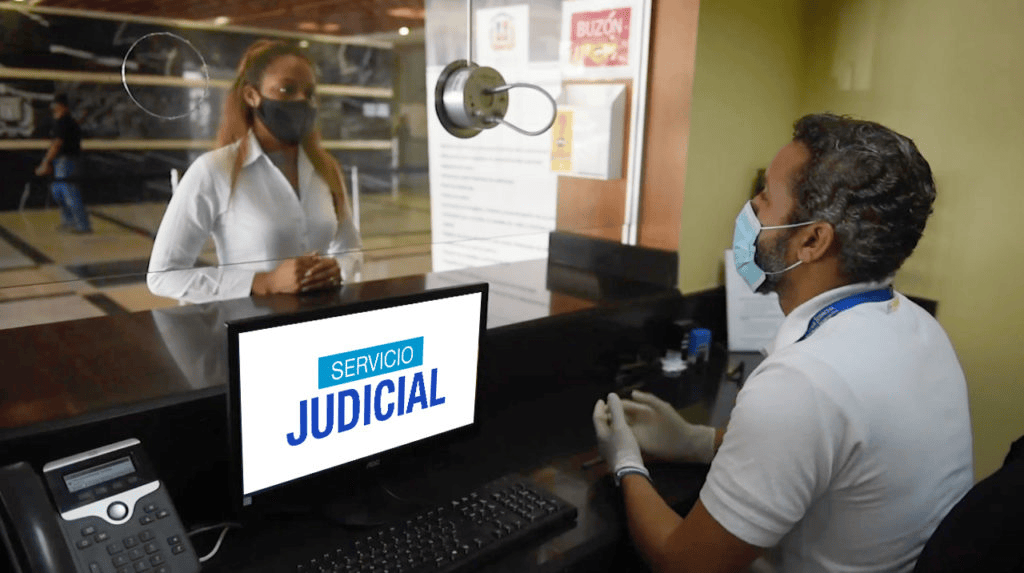 Un hombre con guantes y mascarilla atiende a una mujer en una ventanilla de servicio judicial, con un ordenador visible en el mostrador.