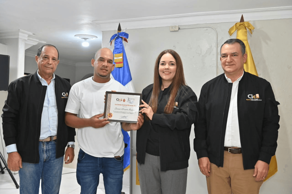 Cuatro personas posando en una ceremonia de entrega de reconocimiento, sosteniendo un certificado. Dos hombres a la izquierda, uno con chaqueta negra y otro con camisa, y dos personas a la derecha, una mujer con chaqueta negra que sostiene el certificado y un hombre con chaqueta blanca.
