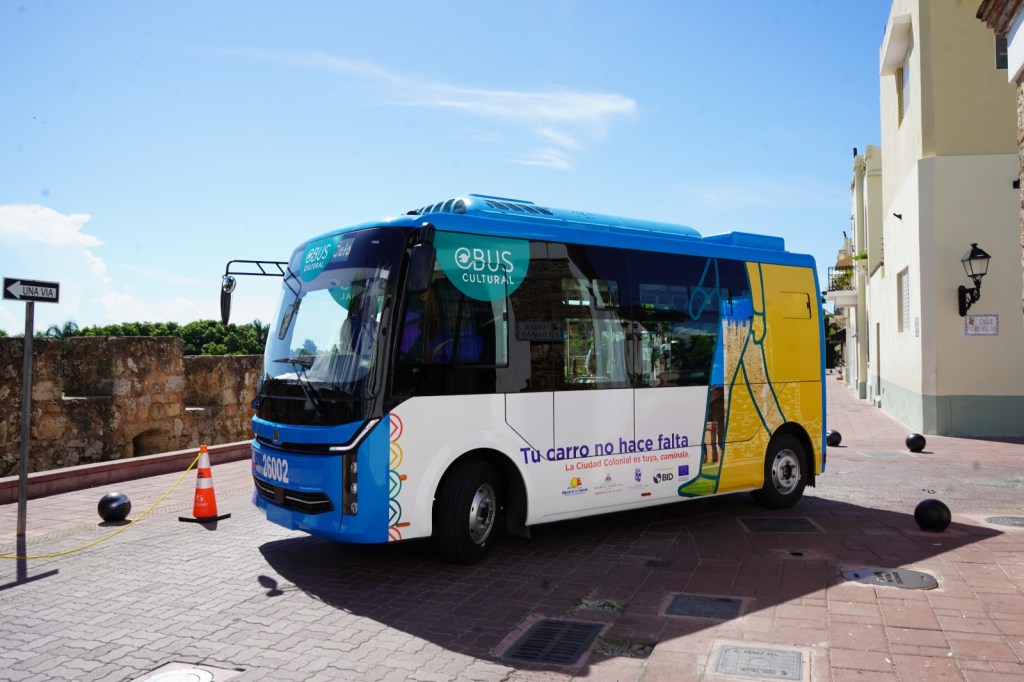 Un autobús cultural colorido estacionado en una calle de la ciudad, con letreros que promueven el uso del transporte público.